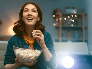 a woman with a happy expression and a projector next to her, eating popcorn, and appearing to be watching a movie