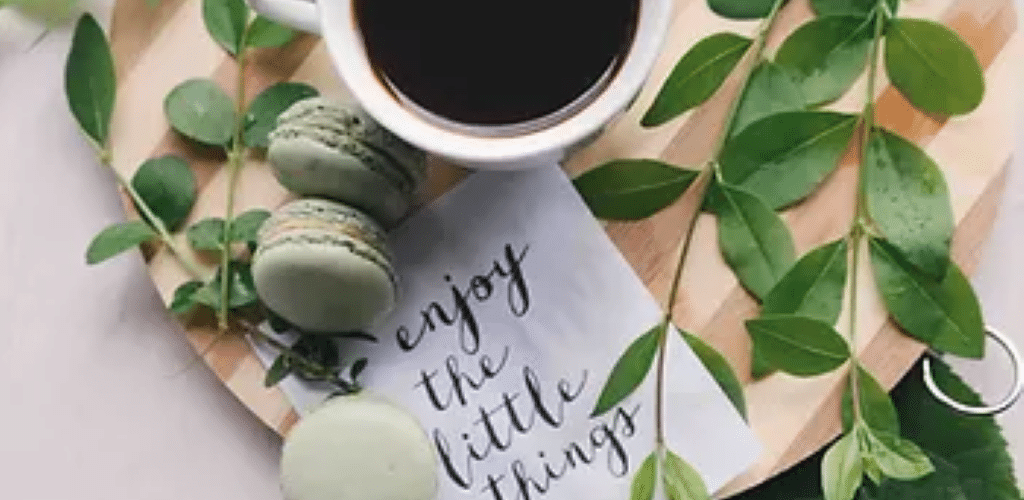 Birds eye image of a cup of coffee on a wooden board, next to some foliage, with green macarons, and a note saying 'enjoy the little things'.