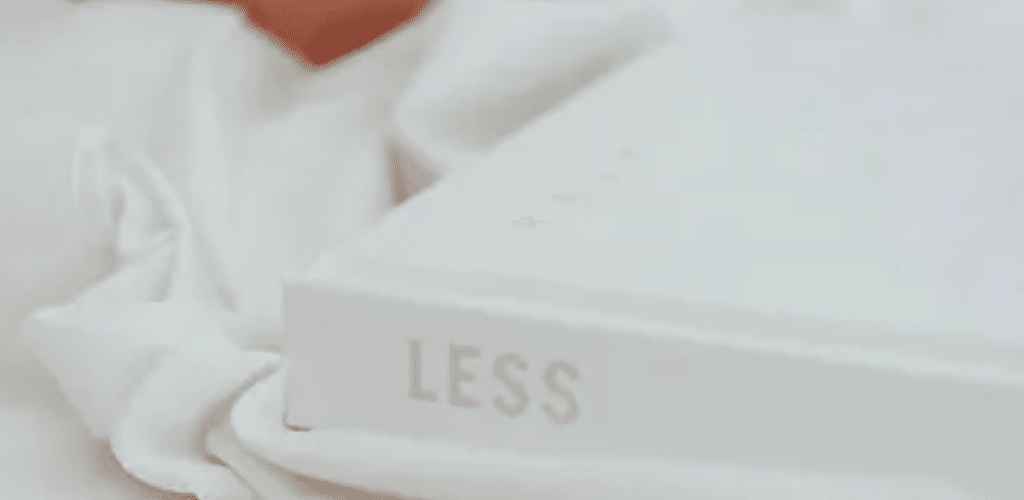 Photo of a white book placed on white sheets or a table cloth with the word 'Less' printed on the book's spine. Close to the book in the background part of a peach-coloured mug can be seen.