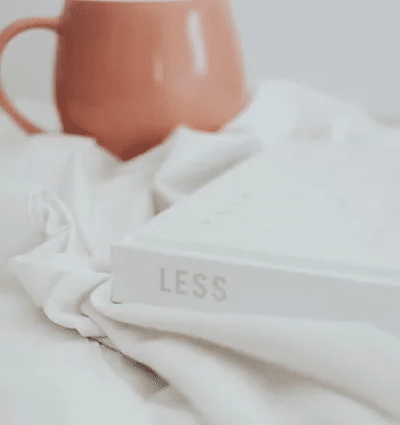 Photo of a white book placed on white sheets or a table cloth with the word 'Less' printed on the book's spine. Close to the book in the background is a peach-coloured mug.